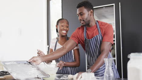 Happy-african-american-couple-in-aprons-baking-together,-preparing-dough-in-kitchen,-slow-motion