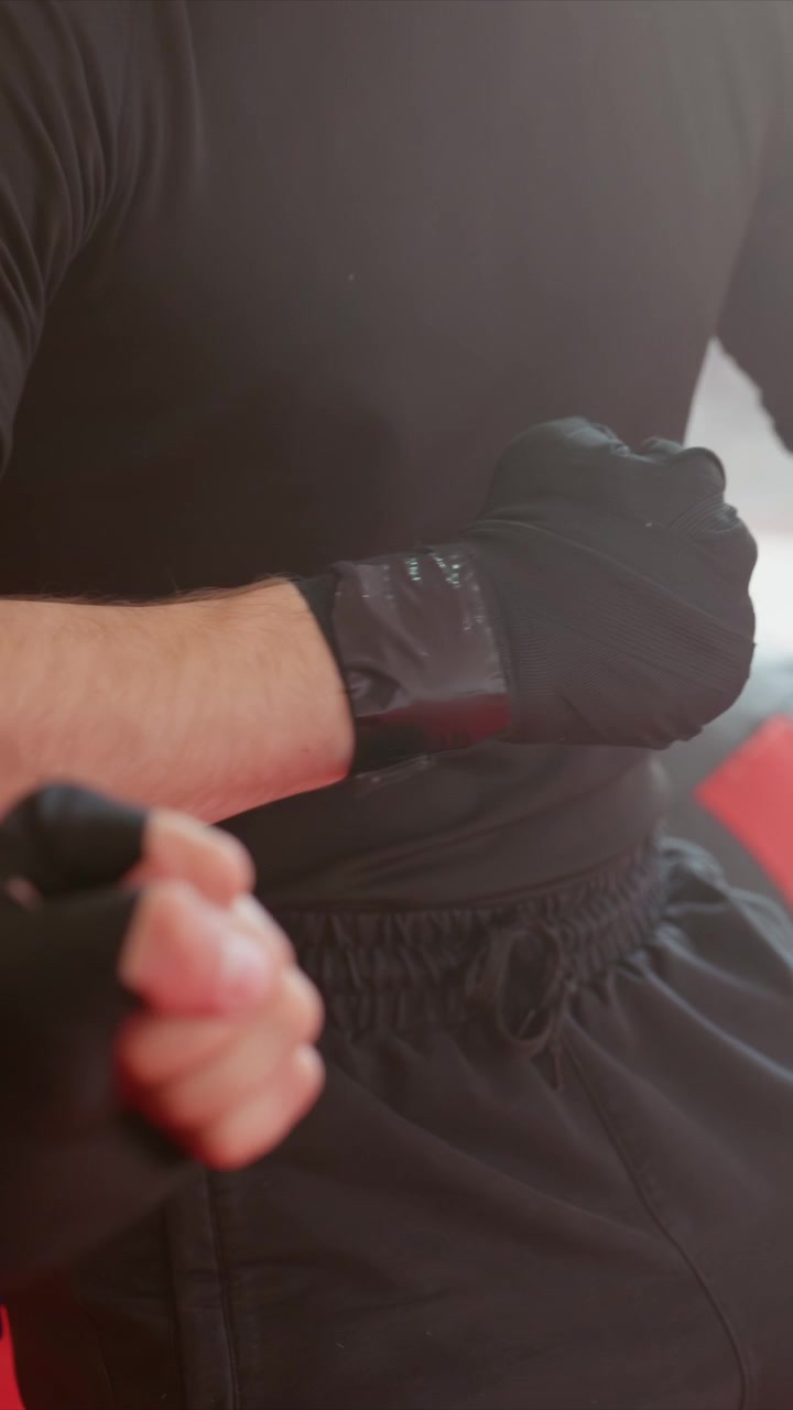 Primer plano de un hombre con ropa negra recibiendo un masaje en el brazo extendido. La escena incluye textura de piel visible y pequeñas marcas. Ambiente interior con suelo de colchoneta roja y pared blanca.
