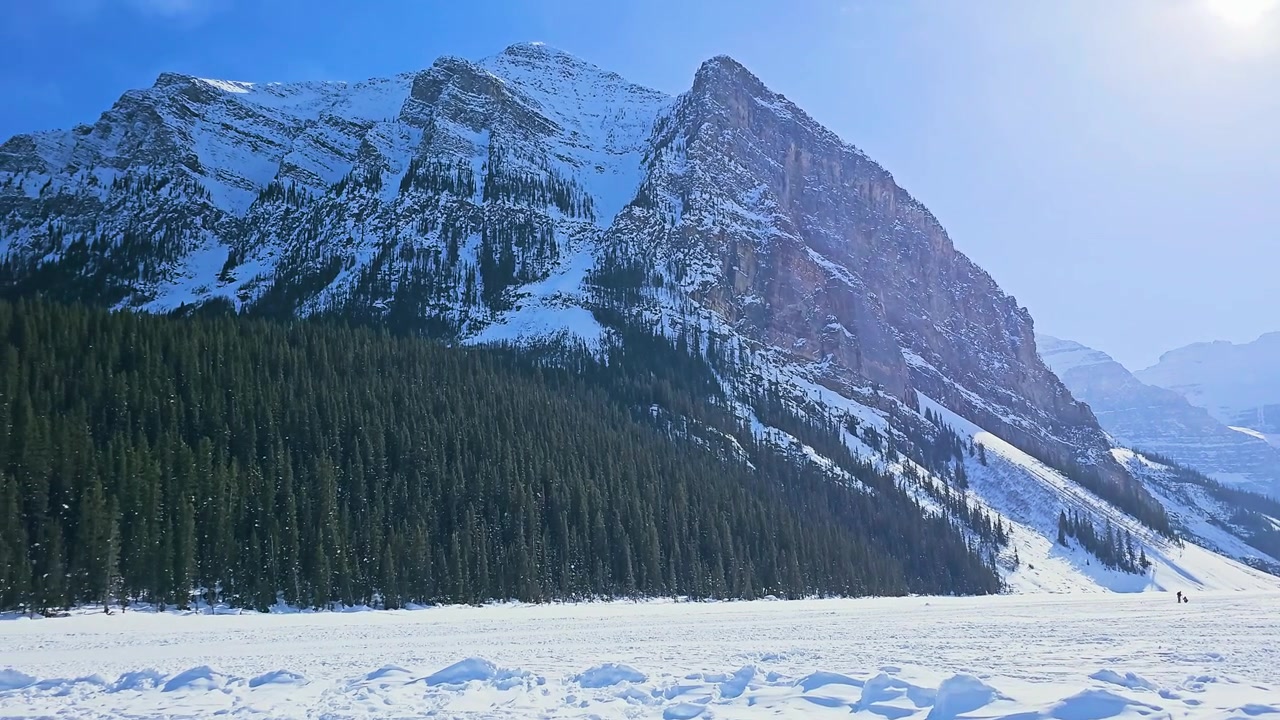 Il paesaggio invernale del Lago Louise nel Parco Nazionale di Banff mostra un lago ghiacciato circondato da aspre cime delle Montagne Rocciose, una fitta foresta di conifere, terreno innevato, cielo luminoso e una luce diurna nitida.