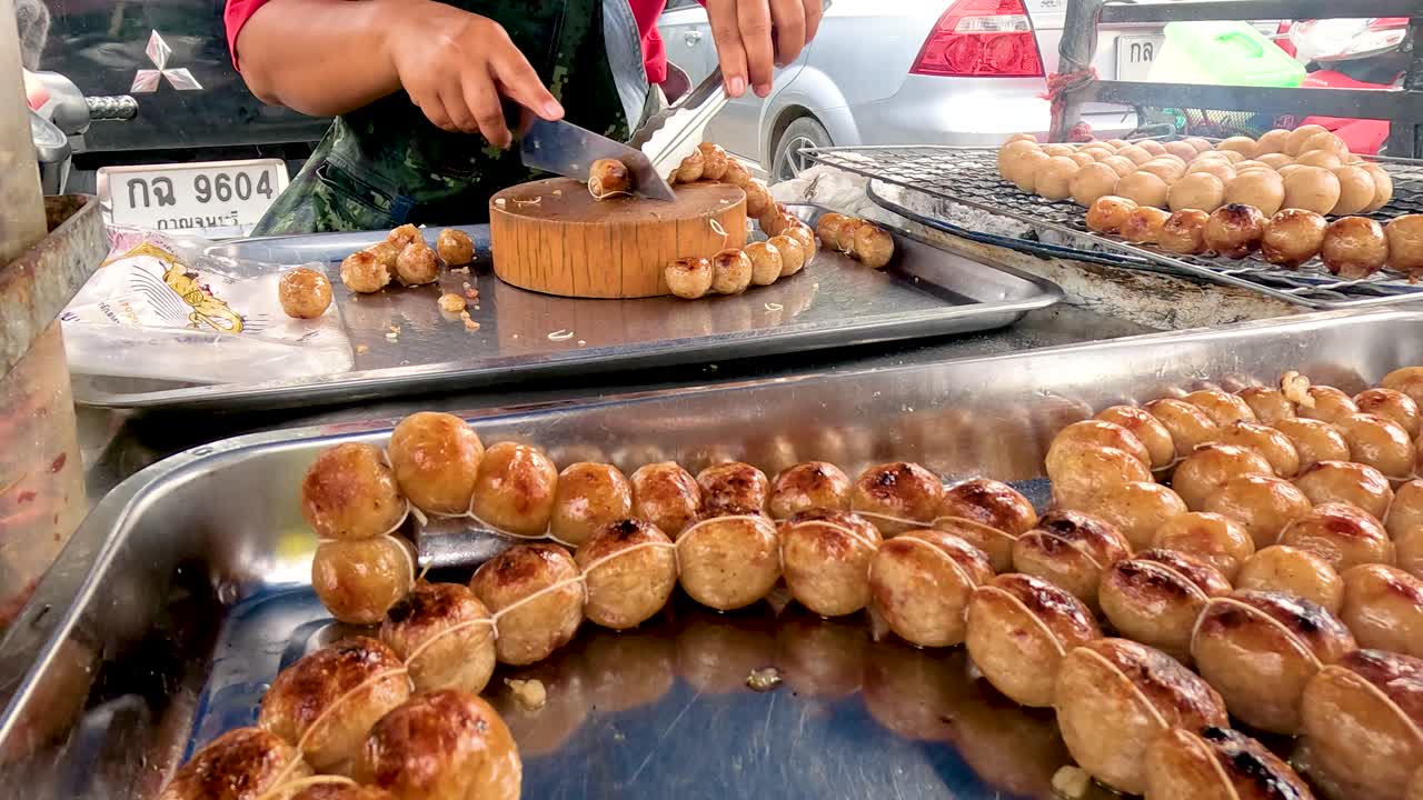 A vendor skillfully prepares grilled sausages at a bustling street market in Kanchanaburi, Thailand, under natural daylight