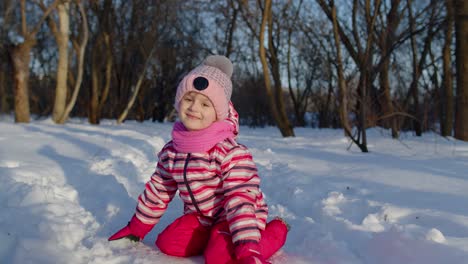 una niña alegre corriendo hacia un camino nevado, mirando a la cámara en el bosque del parque de invierno