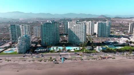 Dolly-En-Vista-Aérea-De-Un-Complejo-De-Edificios-Con-Piscinas-Frente-Al-Mar,-Montañas-áridas-En-El-Horizonte-En-Un-Día-Soleado