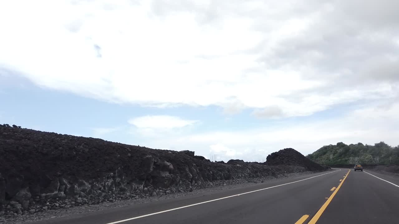 Drive through a lava field on a paved road one year after an eruption with blue skies and clouds.