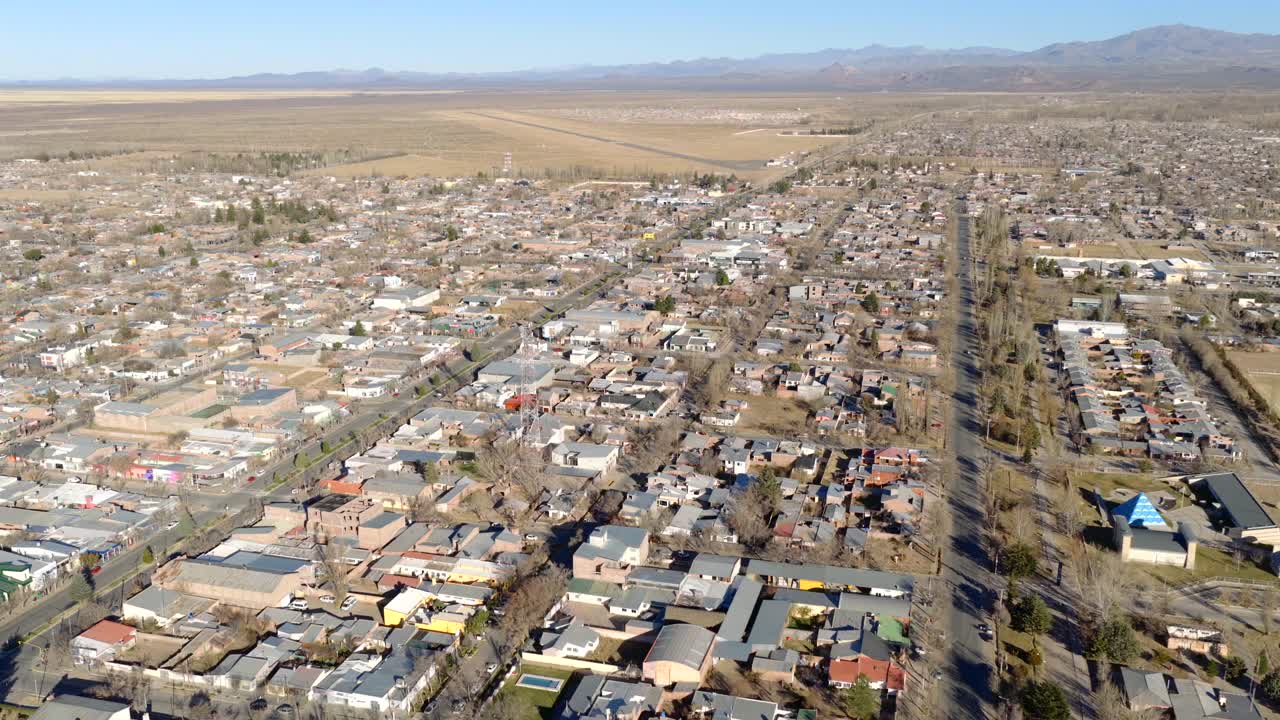 Wide aerial pan over Malargüe, Mendoza, Argentina, showing the town's grid layout, tree-lined streets, low-rise buildings, arid landscape and surrounding mountains under clear blue sky