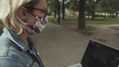 vista lateral de una dama caucásica con anteojos y máscara usando una laptop mientras está sentada en el parque - cerrar