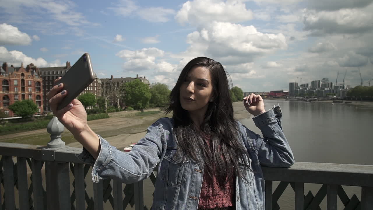 turista latina morena tomando un selfie, posando haciendo el signo de la victoria con la mano, mientras está de pie en la barandilla de un puente en londres