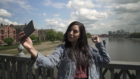 brunette latina tourist taking a selfie, posing doing victory sign with her hand, while standing on the railing of a bridge in london