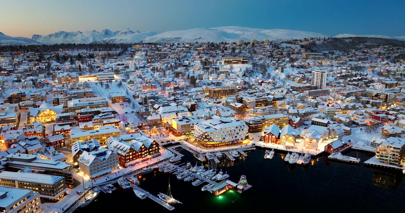 Winterlich schneebedeckte Innenstadt und Hafen von Tromsø mit beleuchteten Straßen. Luftaufnahme.