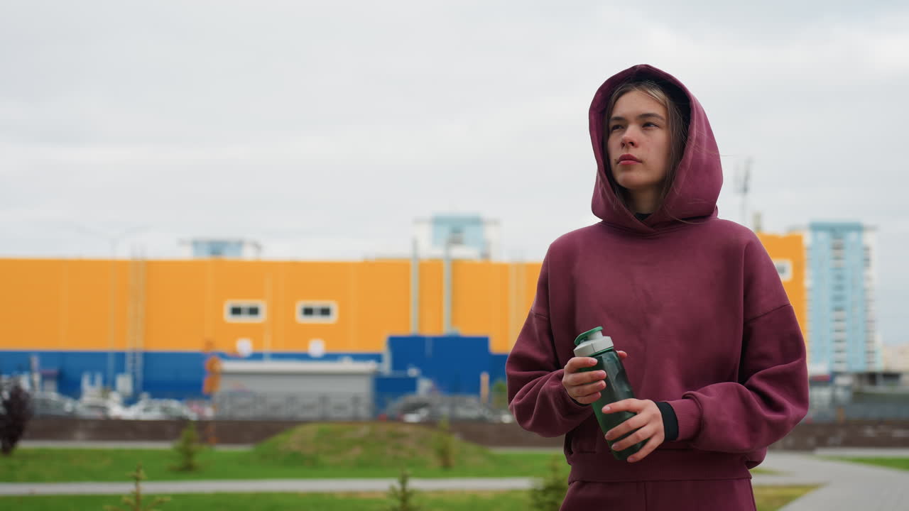 Serene female coach walking along urban pavement clutching water bottle amid spring flowers and high rise apartment buildings under overcast sky showcasing morning wellness lifestyle
