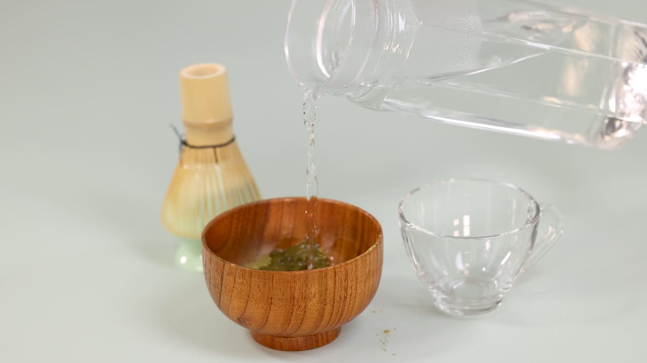 Hot water poured into a wooden bowl with matcha powder, traditional whisk, and cup nearby