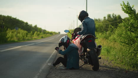 two women by the roadside, one seated on a motorcycle adjusting her clothing while the other crouches down on the ground, closed her helmet visor, both are beside the road