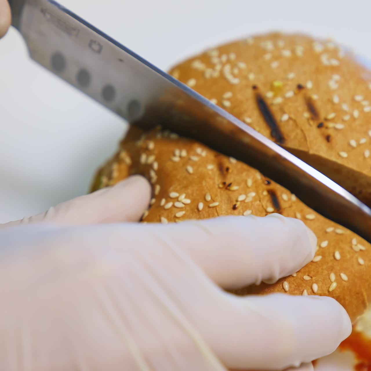 Hands in gloves of a cook making burger. Chef cutting ready hamburger into halves with a sharp knife on table. Close-up