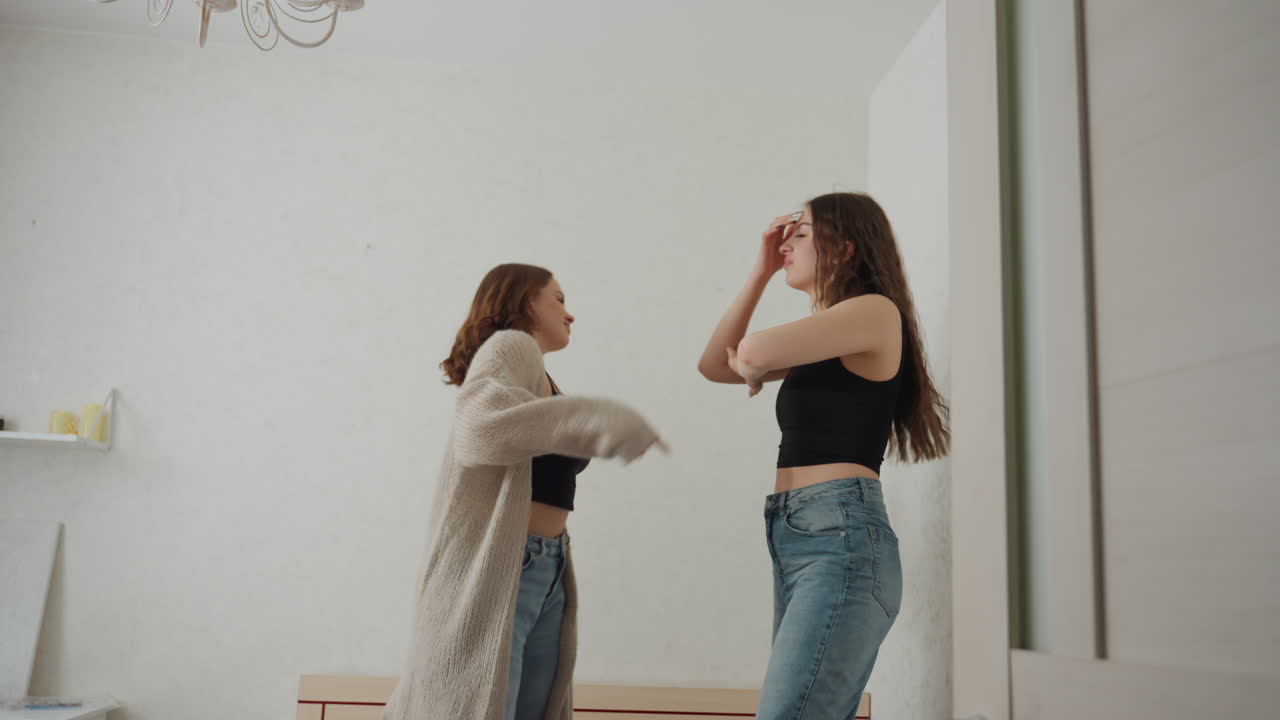 Two Caucasian Friends Styling Together In Front Of Mirror, Casual Jeans And Cardigan Outfits, Playful High Spirits, Bedroom Interior With Chandelier And Wardrobe Shelf, Candid Bonding Moment
