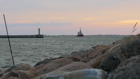 Red-and-white-port-pilot-ship-entering-the-Port-of-Liepaja-in-calm-sunny-summer-evening,-stone-pier-in-foreground,-waves-splashing,-wide-low-angle-shot-from-a-distance