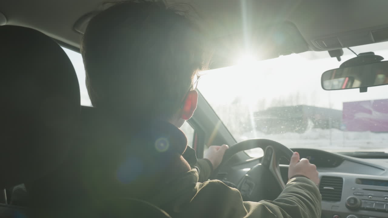 rear view of driver steering car with sunlight filtering through windshield, winter parking lot and blurred buildings in background, hooded jacket silhouette and urban atmosphere with lens flare