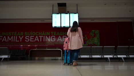 rear shot of a woman with pink raincoat looking at flights panel boarding gate