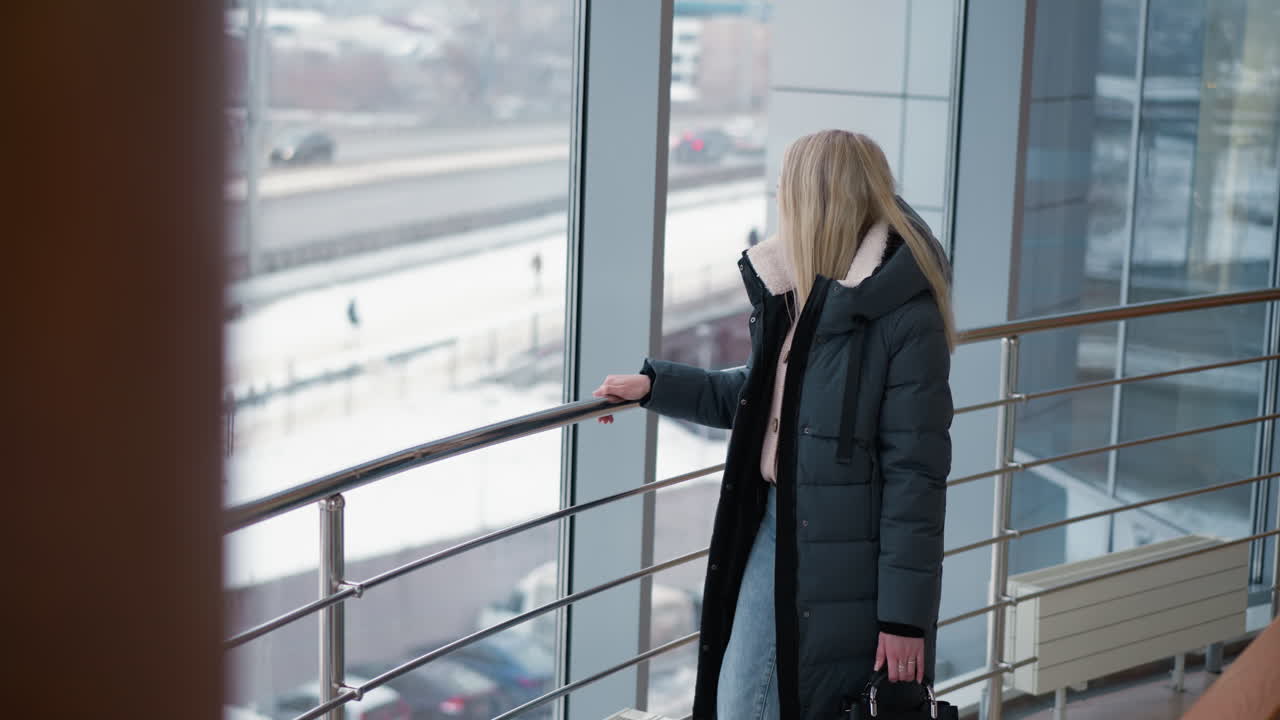 철로를 따라 걸어가는 아름다운 여인, 우아한 금속 레일에 손을 미끄러뜨리고, 세련된 겨울 코트를 입고, 검은 드백을 들고, 유리창을 통해 볼 수 있는 도시 전망과 함께, 도시 환경을 반영합니다.