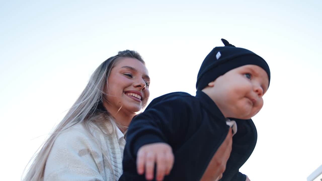 Happy smiling blonde Caucasian woman holding a baby in hands. Mother with her infant child outdoors. Low angle view.