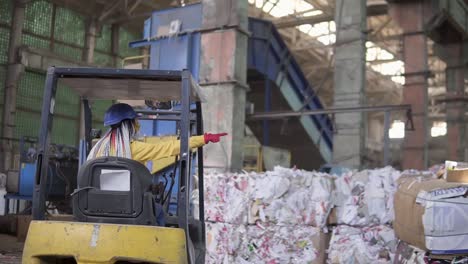 descarga de papel de desecho en un almacén coche eléctrico. trabajadora en casco y chaqueta amarilla sentada en la máquina. enormes existencias de cartón prensado. mujer conductor se sienta desde la parte de atrás y señalando smth