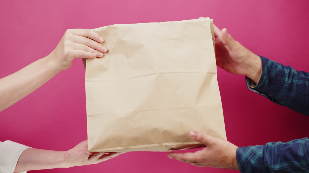 Hands Exchanging a Brown Paper Bag on Pink Background