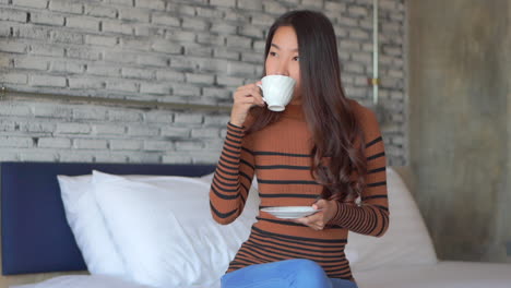 young asian woman drinking morning coffee smiling while sitting on a bed hotel