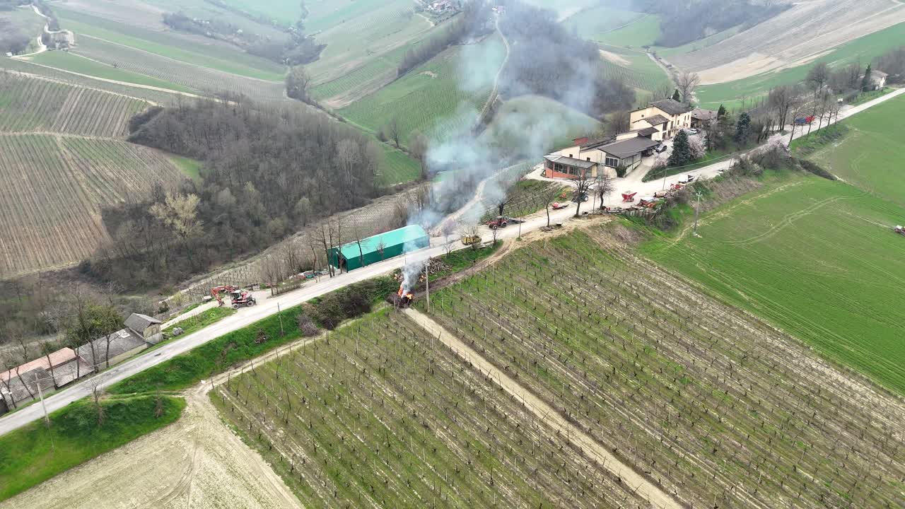 Drone view over Piacenza’s wine valley in Emilia-Romagna, Italy, showing smoke gently rising from burning pruned grape branches along rural roads during late-winter vineyard maintenance