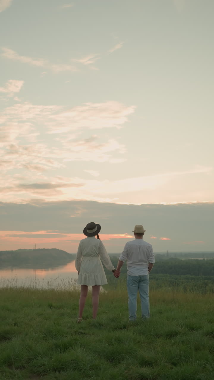 한 부부는 해가 지는 동안 평온한 호수 에 있는 잔디 언덕 위에 서서 하늘을 쳐다보고 있다. 하 셔츠, 모자, 청바지를 입은 남자는 여자의 손을 부드럽게 잡고 있다. 그녀는 검은 모자와  드레스를 입고 있다.