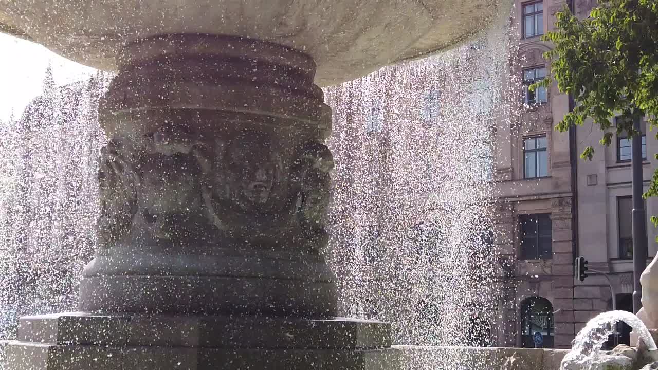 Primer plano a cámara lenta que muestra gotas de agua cayendo de una fuente de piedra ornamentada en una plaza de París. El rocío iluminado por el sol crea un ambiente urbano tranquilo con arquitectura histórica de fondo.