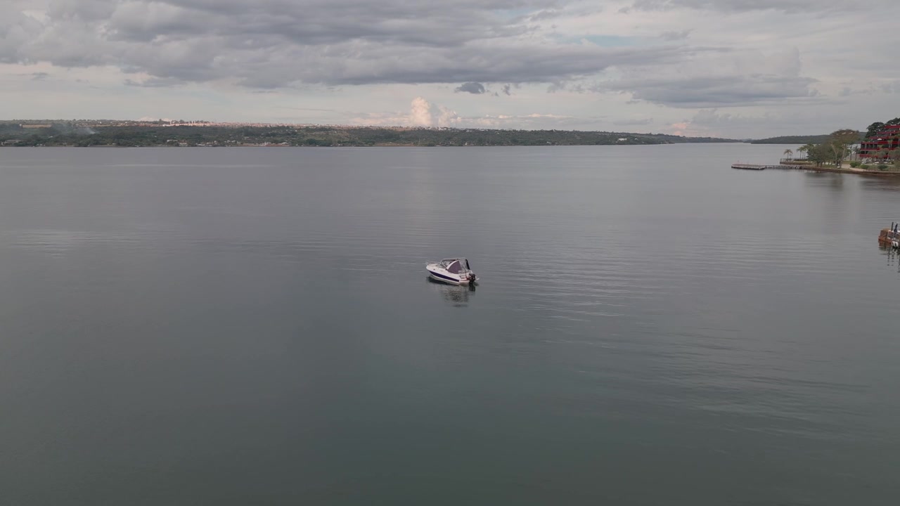 Piccola imbarcazione da diporto che naviga sul Lago Paranoá a Brasilia, Distretto Federale, Brasile, circondata dalle calme acque del bacino, infrastrutture della riva del porto turistico e un'atmosfera tropicale nuvolosa, drone forw