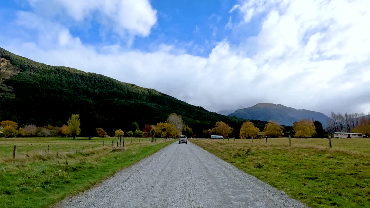 Vehicle travels along rural gravel road, surrounded by autumn trees, mountains, and dynamic sky