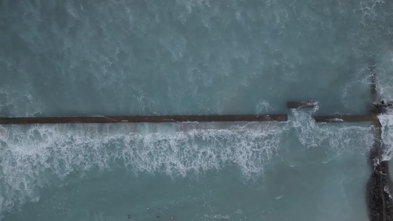 Drone view of ocean waves breaking against a long sea wall, creating white sea foam against deep blue water. The image emphasizes the power of the ocean and human attempts to manage coastal waters
