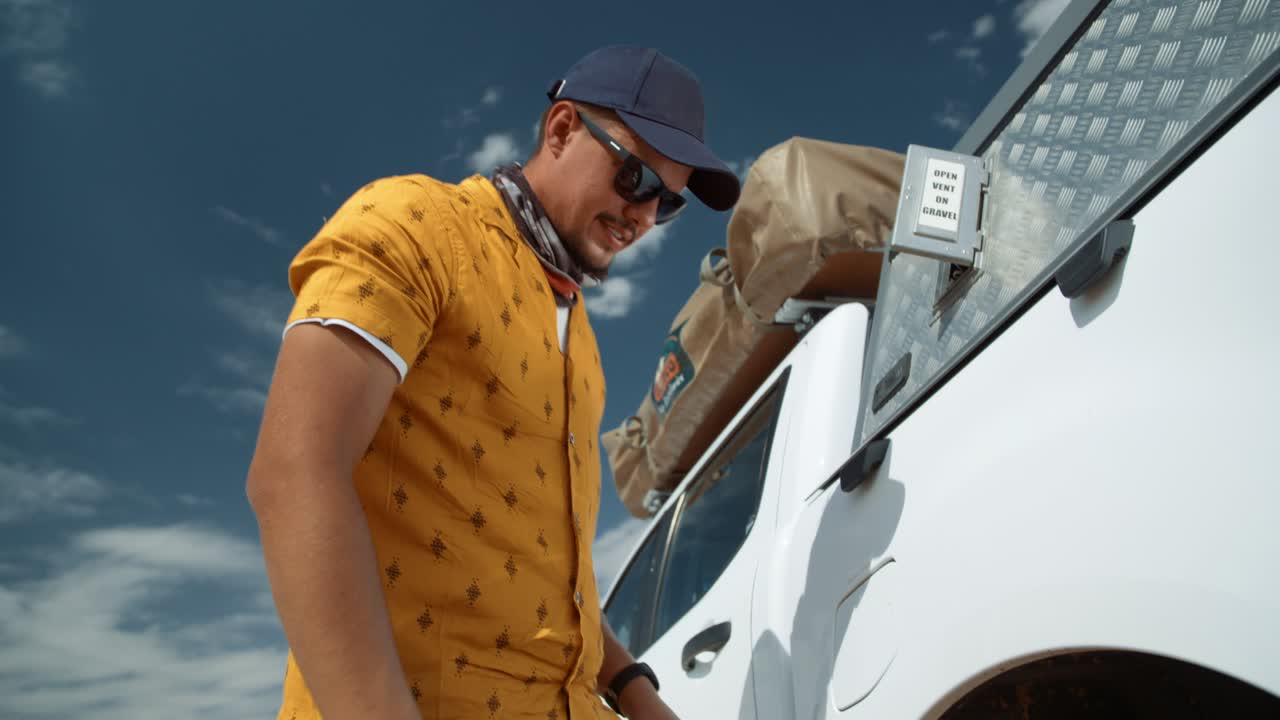 Dynamic tracking low wide hand held shot of a Caucasian male tourist in Africa as he removes a flat tire from the axle of a off-roading vehicle