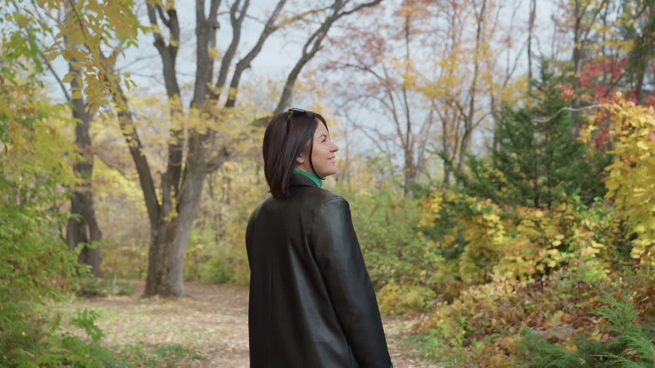 Donna con giacca di pelle che cammina in una foresta con alberi autunnali colorati e cielo nuvoloso; riprese lente da dietro che mostrano dreadlocks e portamento da modella; foglie sotto i piedi e atmosfera tranquilla, cinematografico
