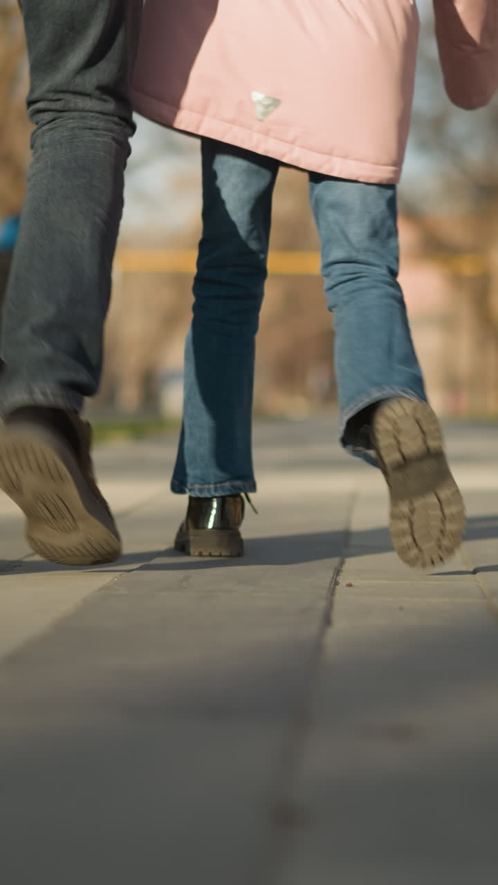 Focused view of a little girl and an adult man's legs walking together on a park pathway, both wearing blue jeans and black shoes in daylight