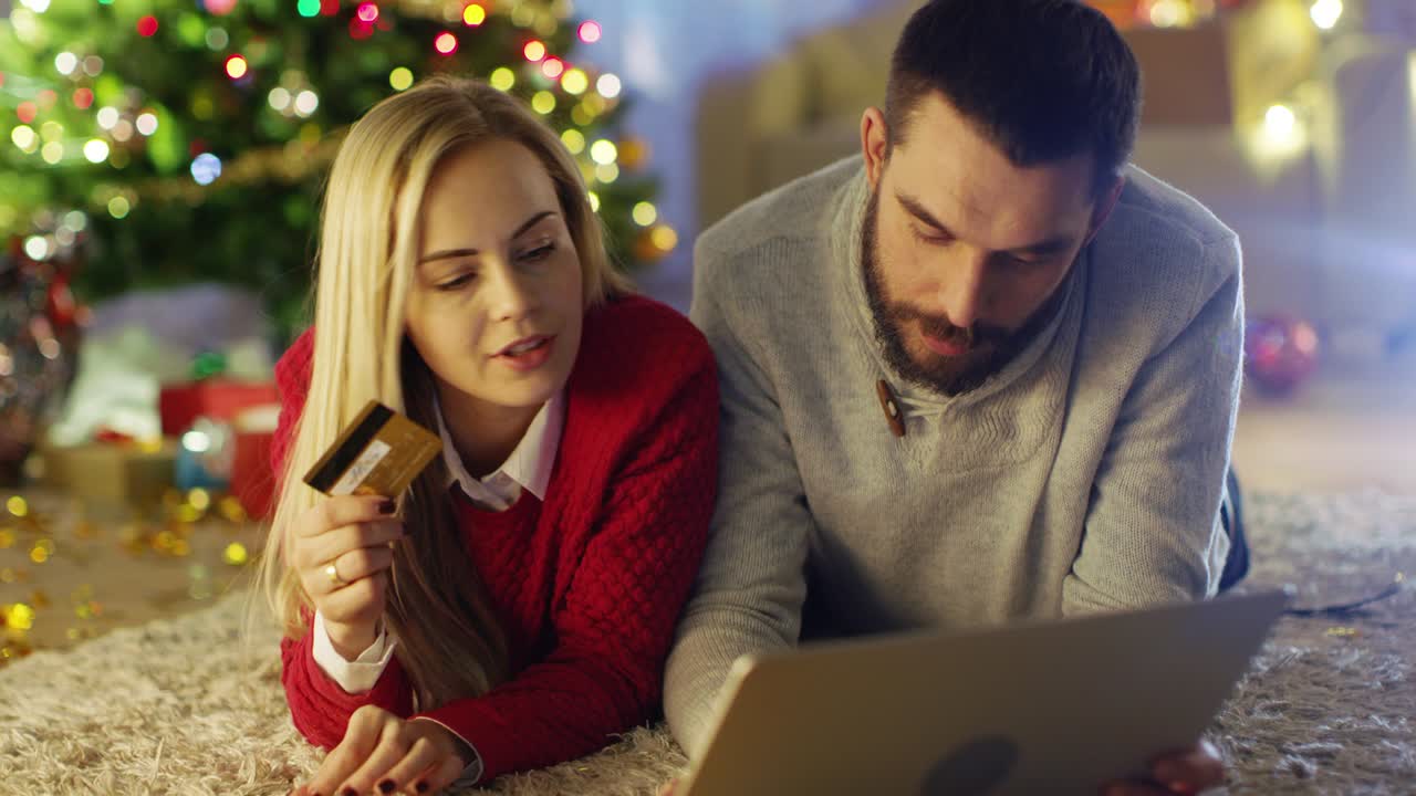 una pareja feliz tendida en la alfombra bajo el árbol de navidad, la mujer tiene una tarjeta de crédito y el hombre le compra regalos en la computadora portátil.