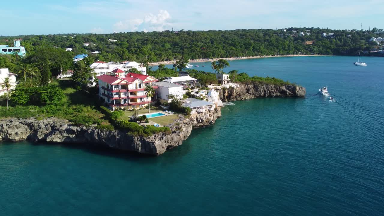 A drone shot of a boat passing by luxurious homes on a cliff side of the north coast of Dominican Republic. In the background you can see Playa Sosua, just outside of the town of Sosua.