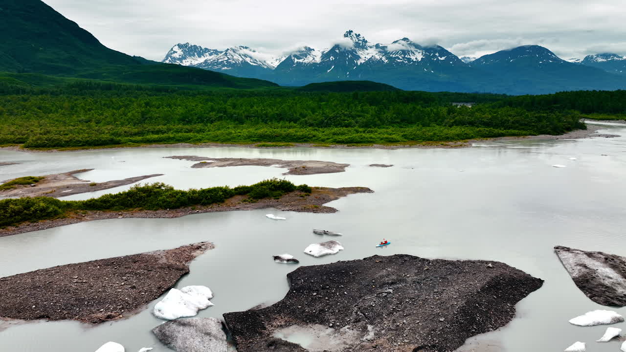 Two people rowing in canoe by the grey river among the boulders of ice. Wooded green waterfront and snow-capped rocks with clouds at tops. Alaska, USA