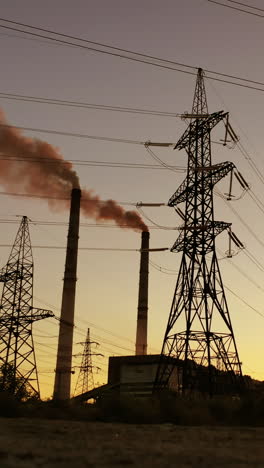 high voltage electric lines and industry. pipes with dark smoke near the transmission electric towers at dusk. pollution the atmosphere. view from below. vertical video