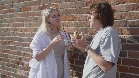 young couple eating a hamburger and sharing a cold drink, while chatting leaning against a brick wall