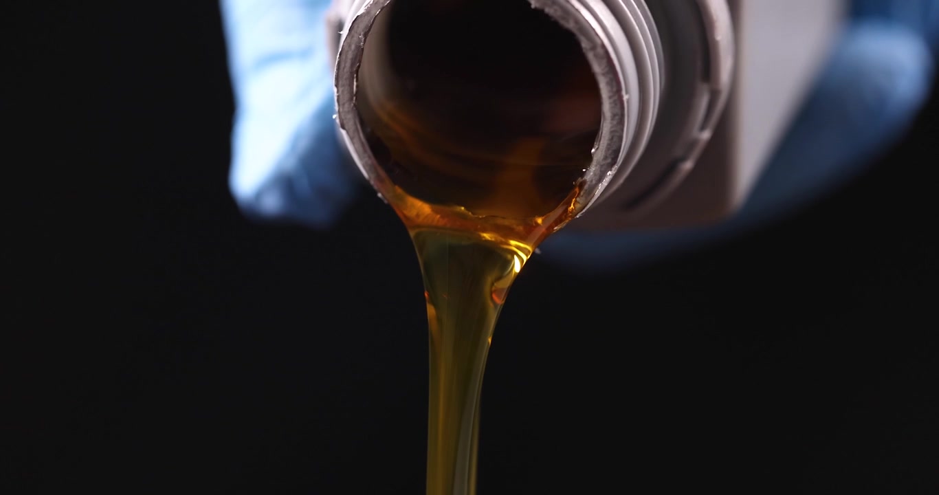 Una persona versa un flusso di olio motore marrone dorato da un contenitore.