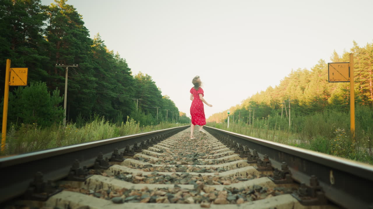 Back view of fair skinned woman in red dress running on railway track through forest and turning head to look back while surrounded by lush greenery and soft evening sunlight in countryside