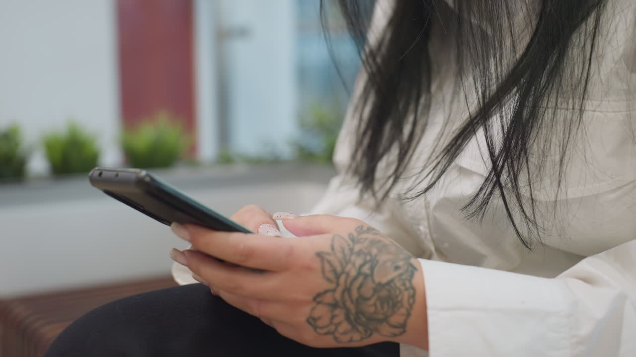 Partial view of woman with floral tattoo on hand using smartphone while seated, surrounded by blurred decorative green plants in background, dressed in white shirt and black pants