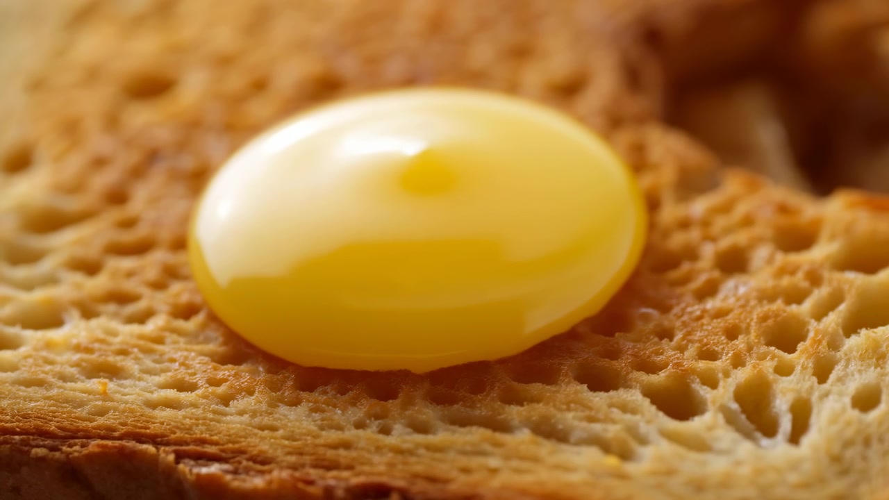 Vista macro dal basso di una salsa gialla lucida che si versa su pane tostato, depositandosi poi in una pozzanghera liscia e arrotondata con una piccola punta centrale, rivelando una calda luce da studio, una crosta croccante e un aspetto appetitoso.