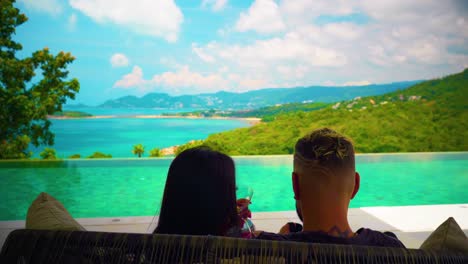 a couple of man and woman on their honeymoon sitting on a sofa by the swimming pool drinking and looking at the sea