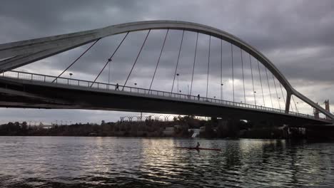 Person-in-River-Kayak-Paddles-under-Bridge-with-Stormy-Clouds-in-Sky