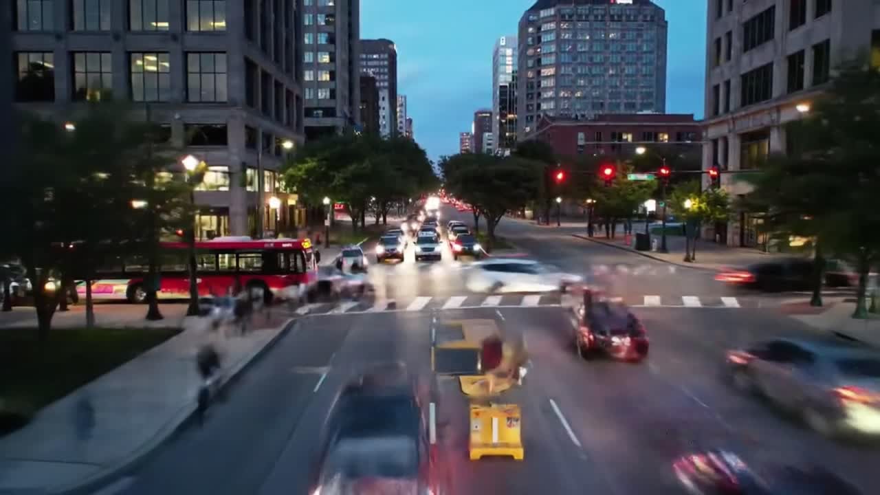 The vibrant atmosphere of a bustling city as cars navigate the streets and pedestrians cross the crosswalks during dusk. The lights from buildings and vehicles create a lively urban scene.