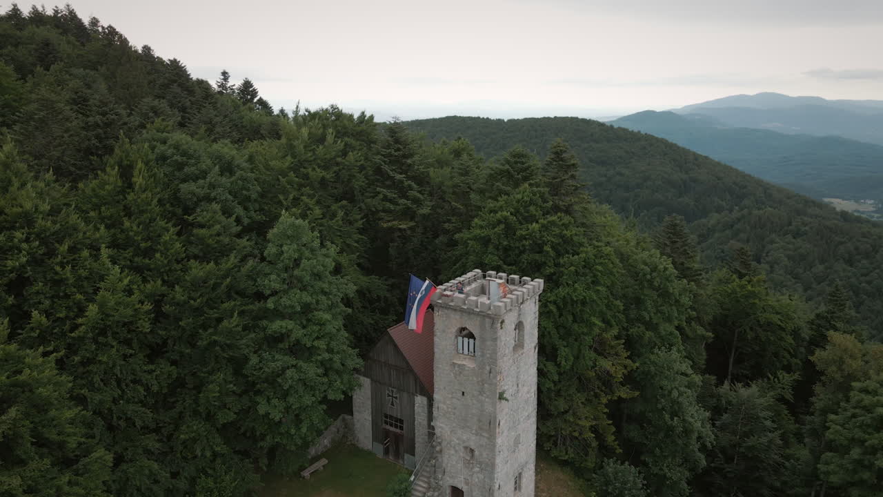 toma de drone de la torre en la montaña mirna gora, joven excursionista de pie en la cima de la torre tomando una vista, de pie cerca de la bandera eslovena y europea