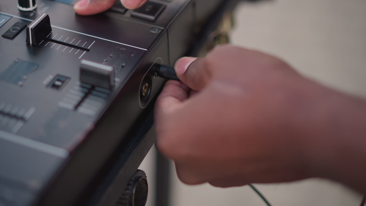 black DJ plugs cable into mixer closeup hands connecting headphones and adjusting fader, urban street setup, warm natural light, focused preparation for live set, tactile movement, professional