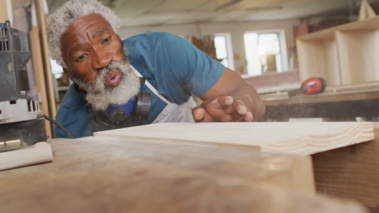 carpintero afroamericano soplando el polvo sobre una tabla de madera en una carpintería