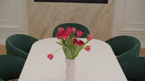 a white vase holding a bouquet of fresh, bright red tulips stands on a sleek white table, with plush green velvet chairs in the background